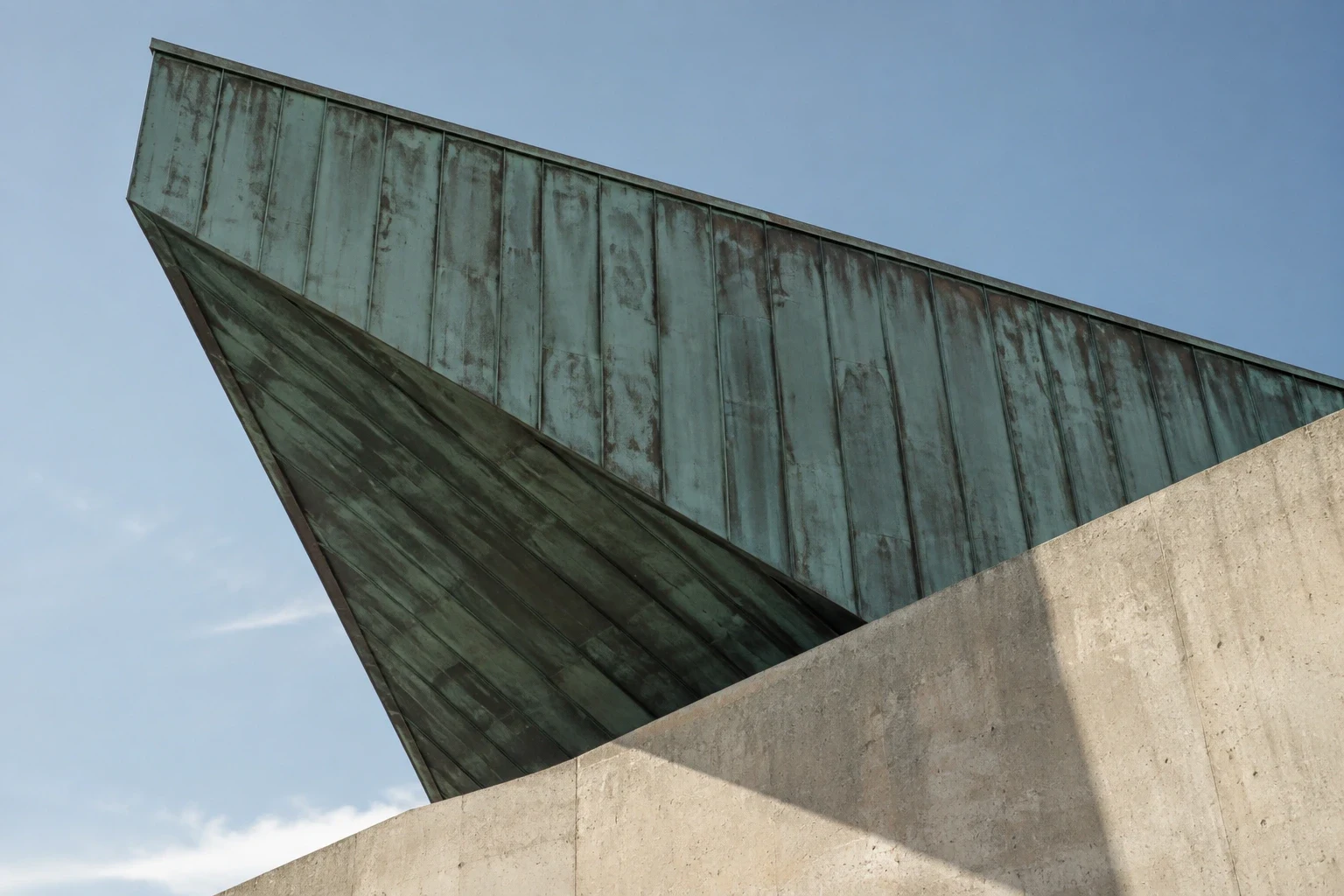 Angular patinated copper roof above a pale concrete wall.